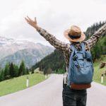 happy-male-traveler-posing-with-hands-up-standing-highway-looking-distant-trees