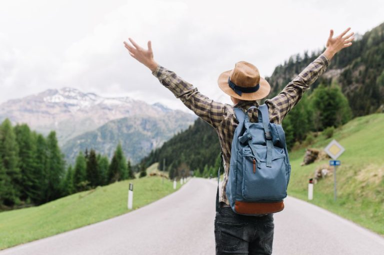 happy-male-traveler-posing-with-hands-up-standing-highway-looking-distant-trees