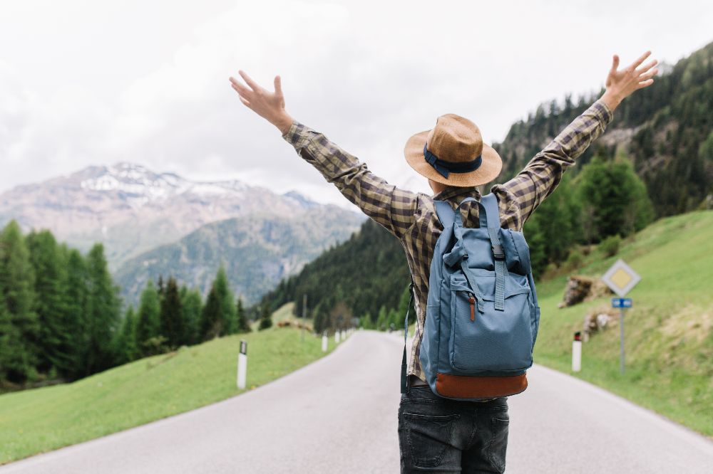 happy-male-traveler-posing-with-hands-up-standing-highway-looking-distant-trees